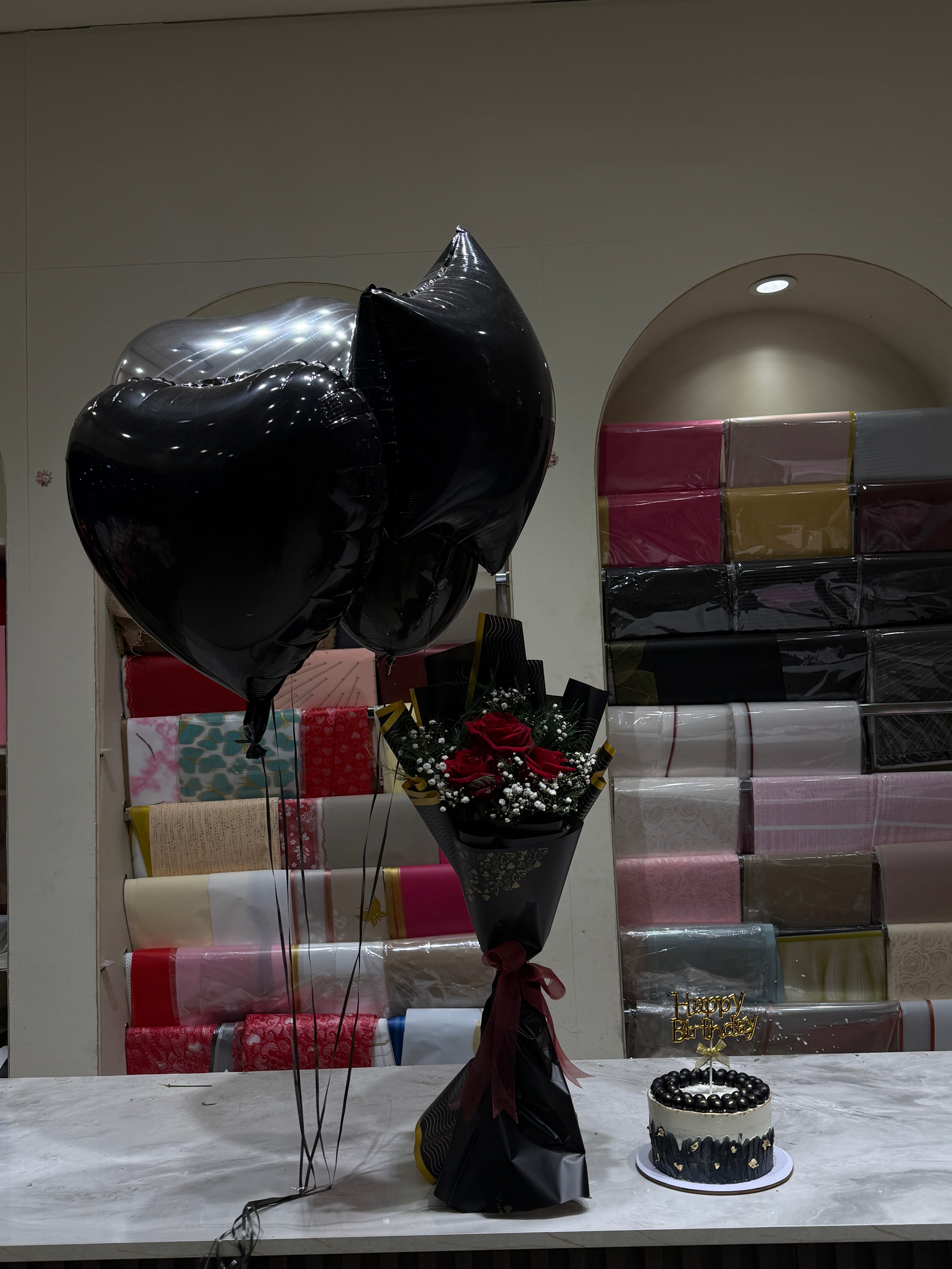 A black heart-shaped balloon and a black-wrapped bouquet of red roses with white filler flowers, displayed on a table next to a small black and white cake in front of a gift shop background with wrapped presents.