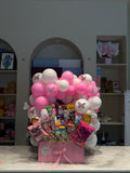 A pink Mother's Day gift basket filled with assorted chocolates, candies, and small flowers, decorated with pink and white balloons and butterfly stickers, set on a marble countertop in front of a gray wall.