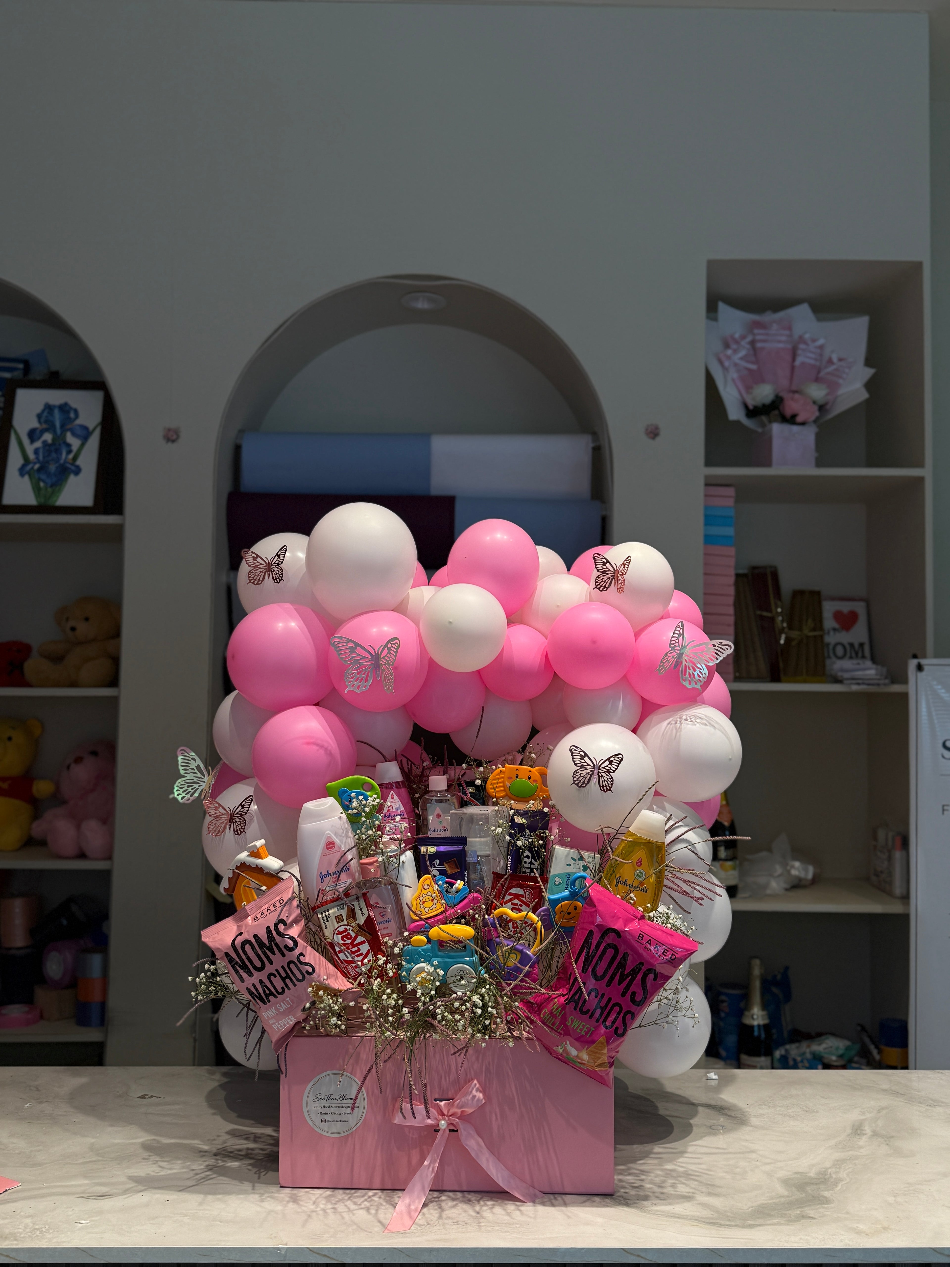 A pink Mother's Day gift basket filled with assorted chocolates, candies, and small flowers, decorated with pink and white balloons and butterfly stickers, set on a marble countertop in front of a gray wall.