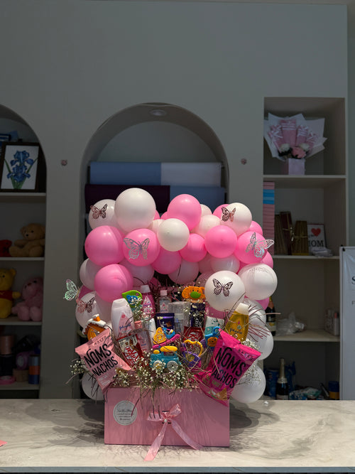 A pink Mother's Day gift basket filled with assorted chocolates, candies, and small flowers, decorated with pink and white balloons and butterfly stickers, set on a marble countertop in front of a gray wall.