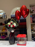 A gift arrangement on a table featuring a black-wrapped flower bouquet with red and white roses, three metallic red heart-shaped balloons, and a small cake in a clear red gift box.