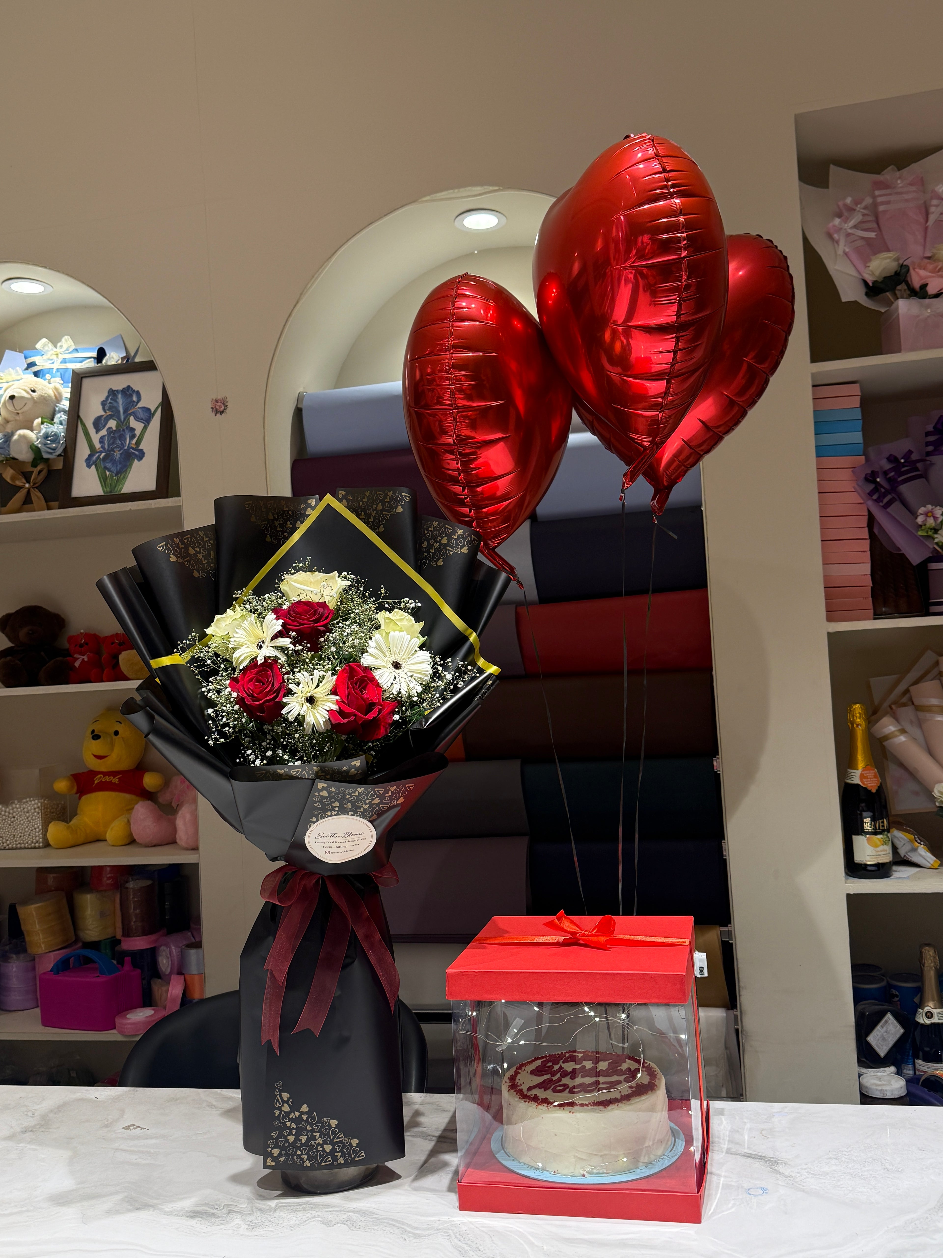 A gift arrangement on a table featuring a black-wrapped flower bouquet with red and white roses, three metallic red heart-shaped balloons, and a small cake in a clear red gift box.