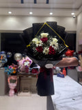 A hand holding a wrapped bouquet of red and white roses with white daisies, baby's breath, and green foliage, wrapped in black paper with a gold accent line and a dark red ribbon, in a shop setting.