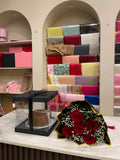 A chocolate cake in a clear square gift box with a black lid and ribbon, placed on a marble counter next to a bouquet of red roses with white filler, in front of shelves filled with wrapped gift boxes and wrapping paper rolls.