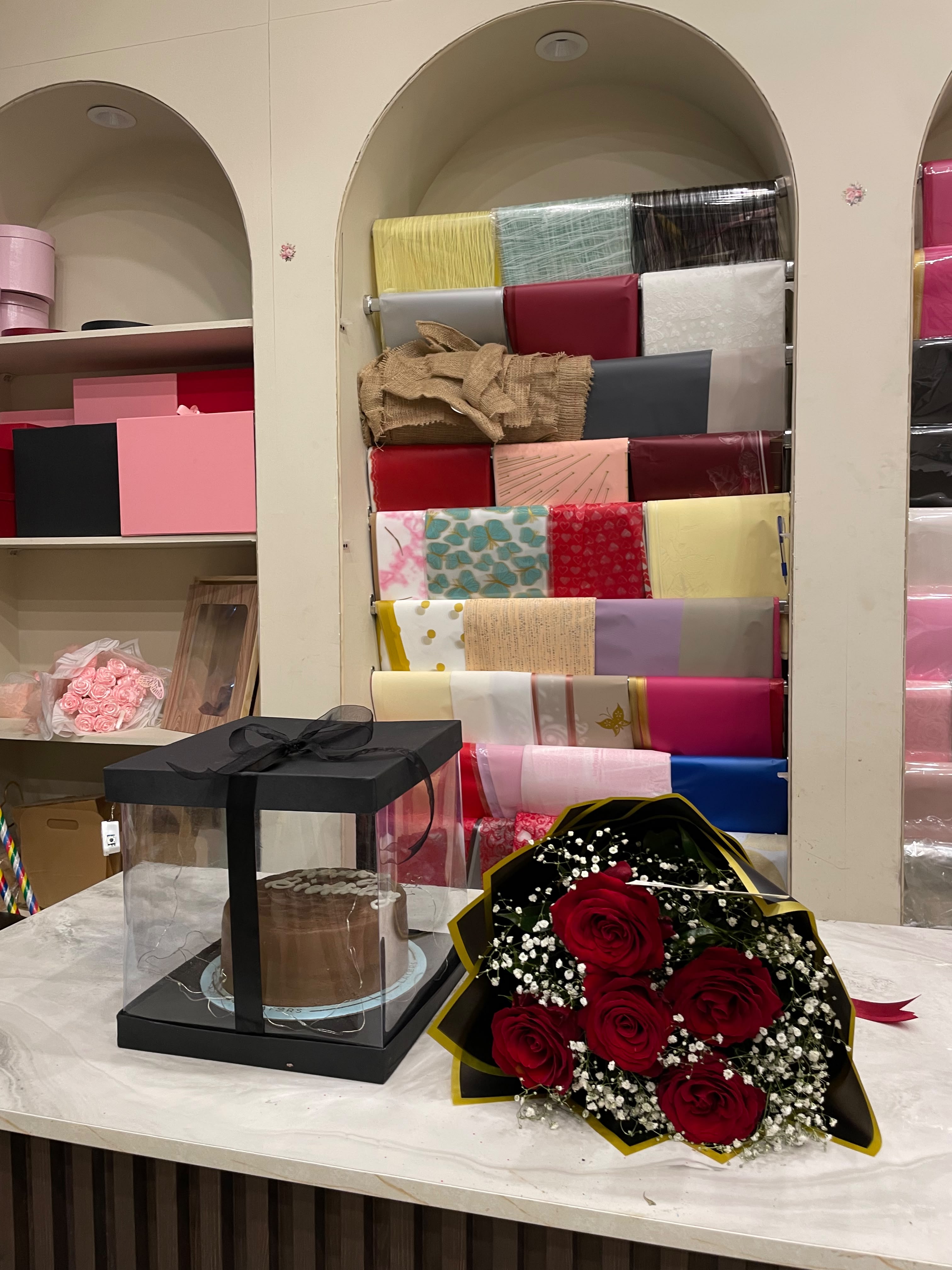 A chocolate cake in a clear square gift box with a black lid and ribbon, placed on a marble counter next to a bouquet of red roses with white filler, in front of shelves filled with wrapped gift boxes and wrapping paper rolls.