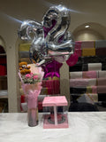 A birthday gift arrangement featuring a large metallic silver number 2 foil balloon, a pink flower bouquet in a clear vase with a teddy bear, and a pink tiered cake in a clear box on a marble countertop.