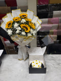 A bouquet of sunflowers and white flowers wrapped in white paper with gold trim sits on a table next to a small round cake in an open black box. The background shows a busy indoor setting with wrapped packages.