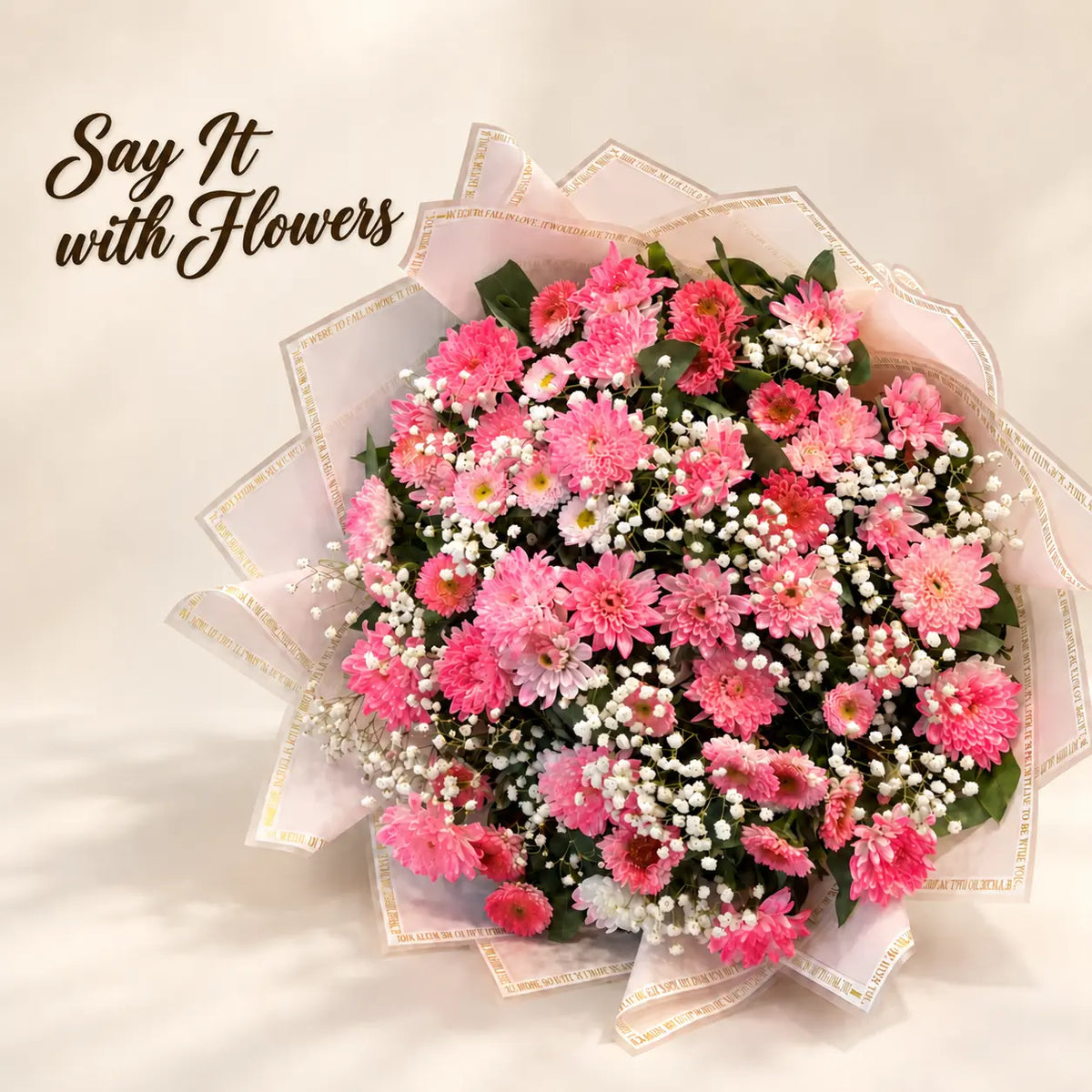 A large floral bouquet filled with pink and red chrysanthemums, white baby's breath, and green foliage, wrapped in sheer white paper with gold trim. The bouquet is set against a warm beige background with the text 'Say It with Flowers' in the upper left corner.