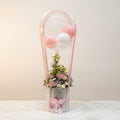 Decorative flower arrangement with balloons in a clear holder on a textured surface.