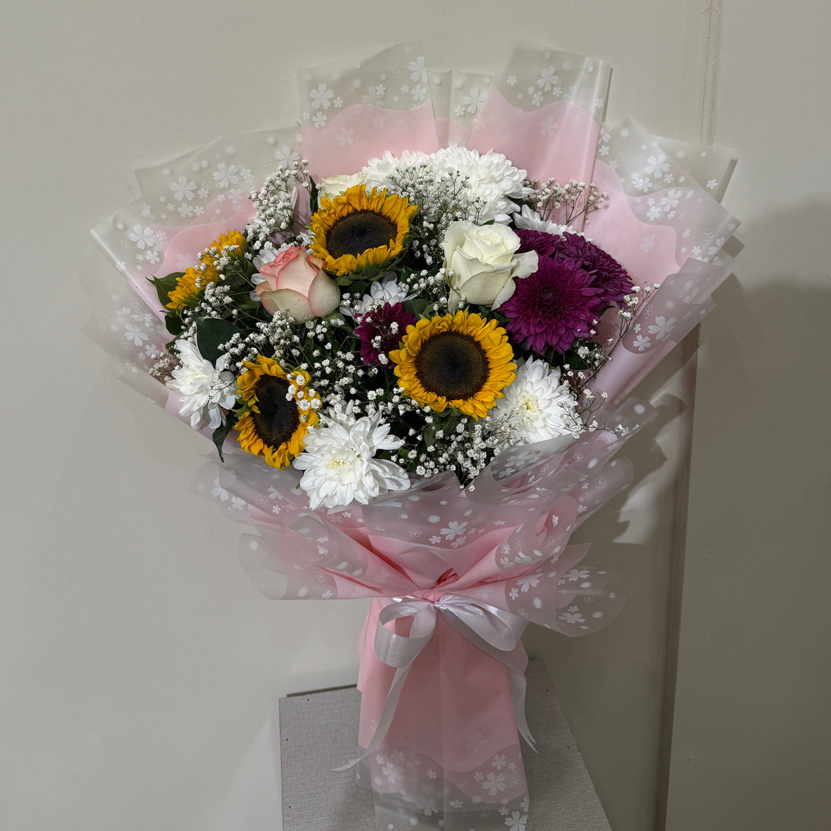 A mixed flower bouquet wrapped in pink patterned tissue paper and tied with a pink ribbon, featuring sunflowers, white chrysanthemums, purple flowers, and baby's breath, displayed on a small pedestal against a plain wall.