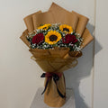 A floral bouquet wrapped in brown kraft paper, featuring yellow sunflowers, red roses, and white baby's breath, tied with a dark ribbon at the base, set against a plain off-white wall.
