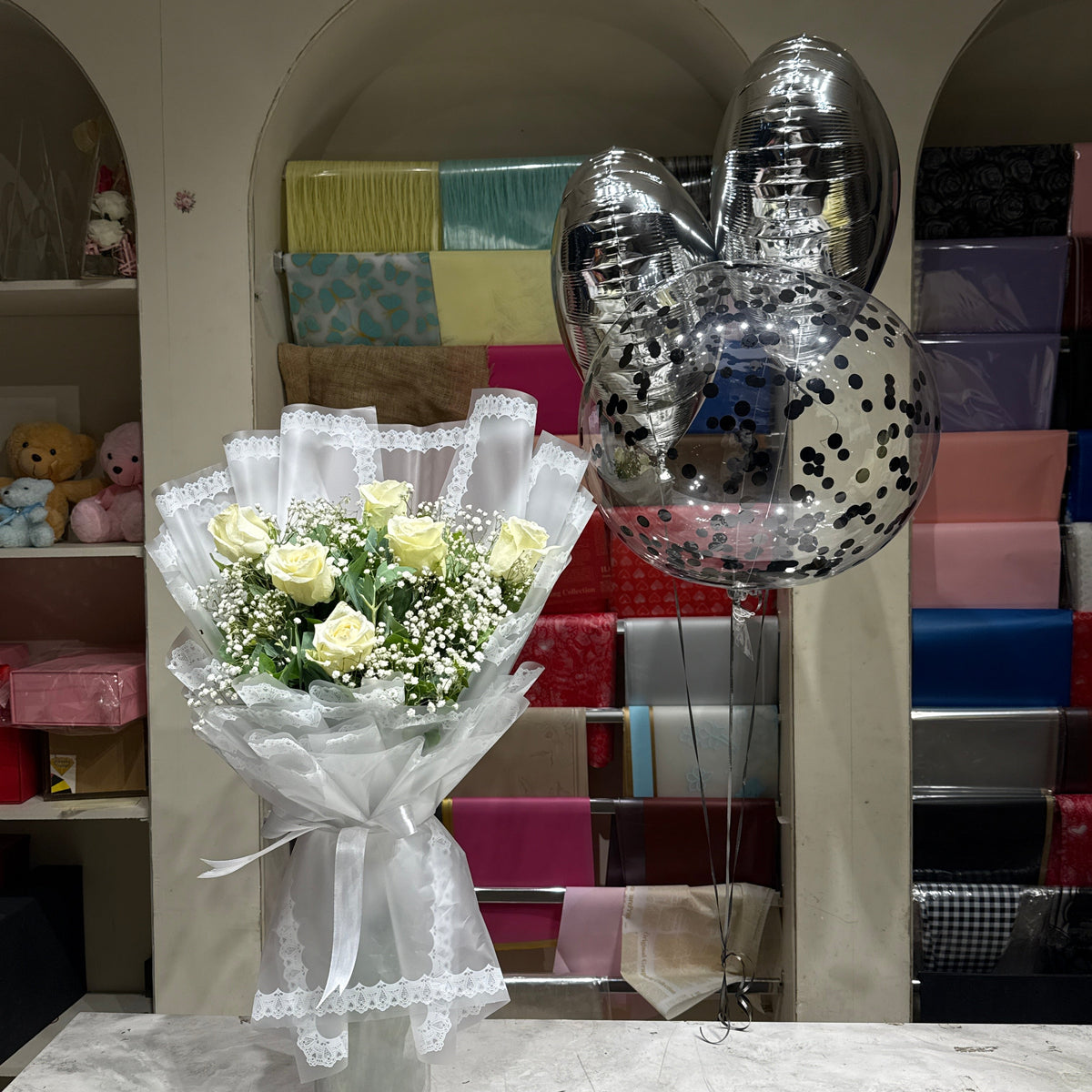 A bouquet of pale yellow roses wrapped in white lace ribbon and sheer white tissue paper, placed on a marble-like surface next to a cluster of metallic silver balloons with black confetti, set against a background of gift boxes in an archway.