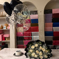 A bouquet of cream roses and baby's breath in black paper wrapping sits on a marble counter next to a small round cake, with black and clear balloons floating in the background against a wall filled with colorful gift boxes.