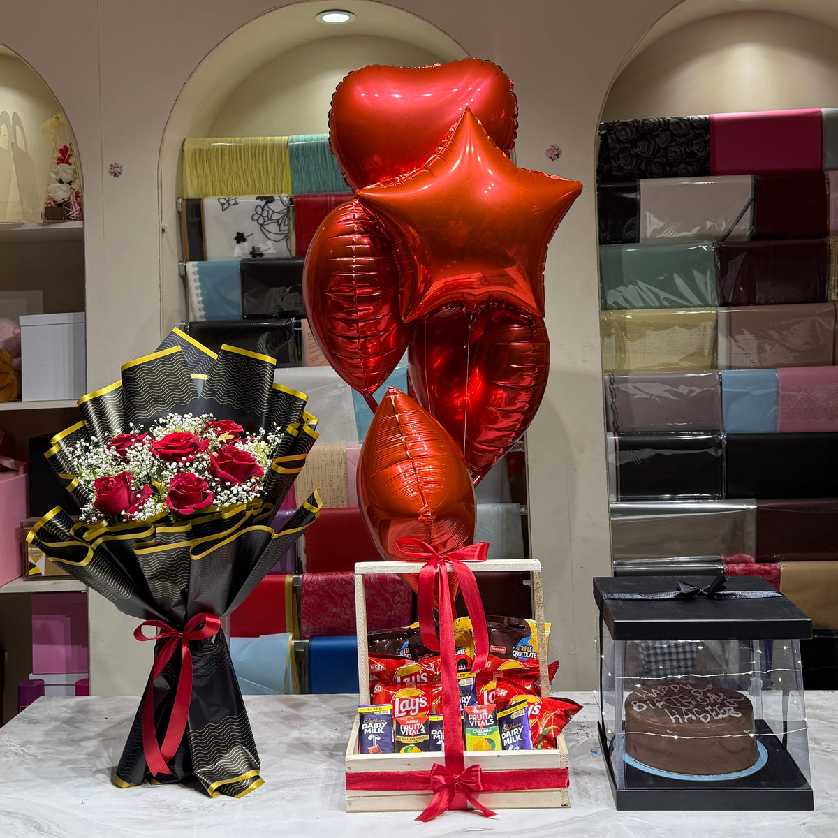 A gift set arrangement on a table featuring a bouquet of red roses in black and gold wrapping, a cluster of metallic red heart and star balloons, a small gift basket with assorted candy, and a chocolate cake under a clear display box, set against a gift shop background.