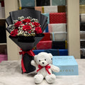 A bouquet of red roses wrapped in black paper with a red ribbon, a small white teddy bear with a red bow, and a light blue gift box on a marble-like surface in front of a gift shop display.