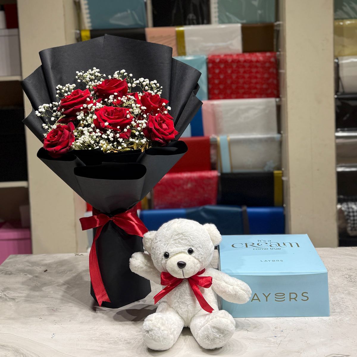 A bouquet of red roses wrapped in black paper with a red ribbon, a small white teddy bear with a red bow, and a light blue gift box on a marble-like surface in front of a gift shop display.