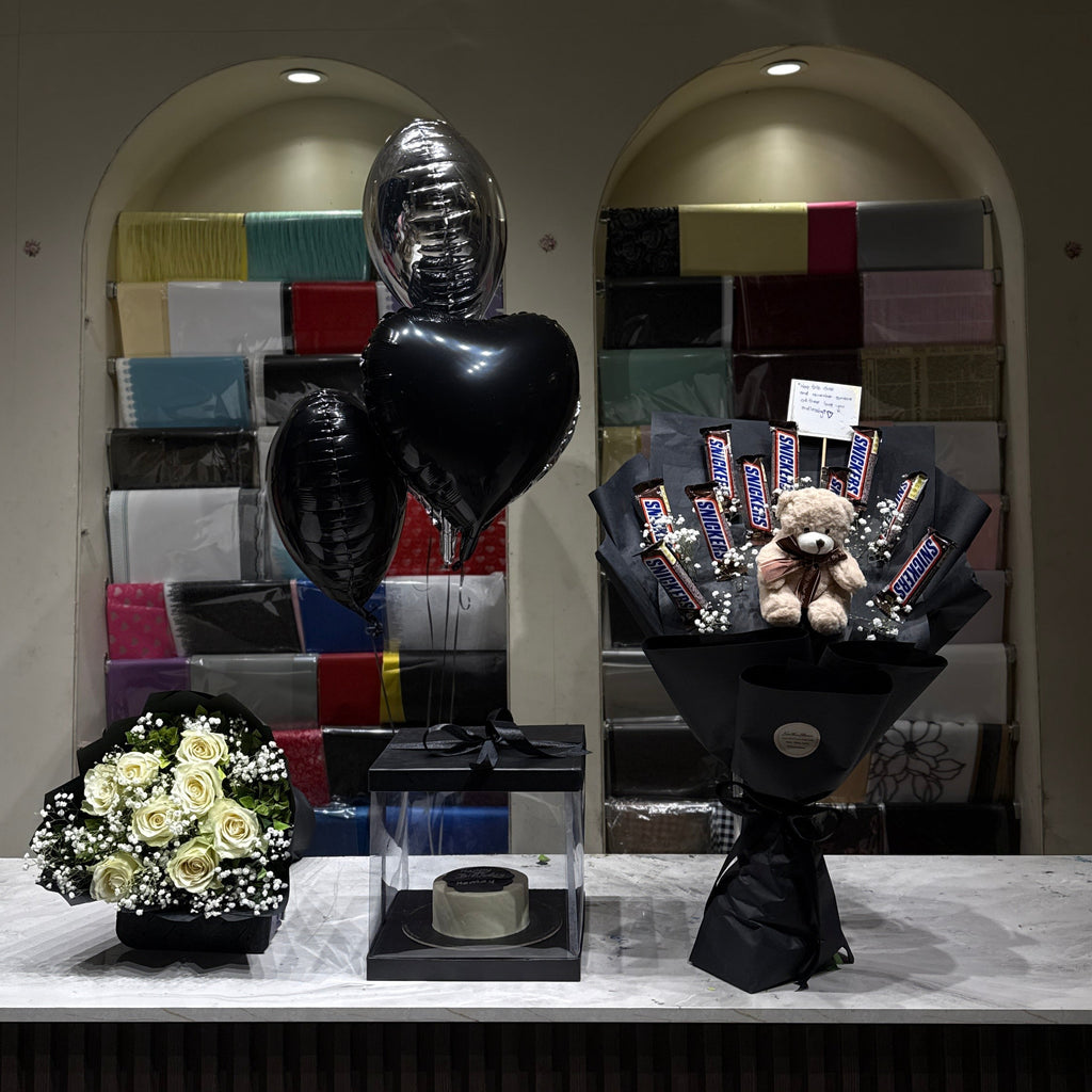 A black memorial balloon bouquet with a heart-shaped balloon and a black flower arrangement with a teddy bear bouquet, displayed on a marble counter in front of a gift shop background.