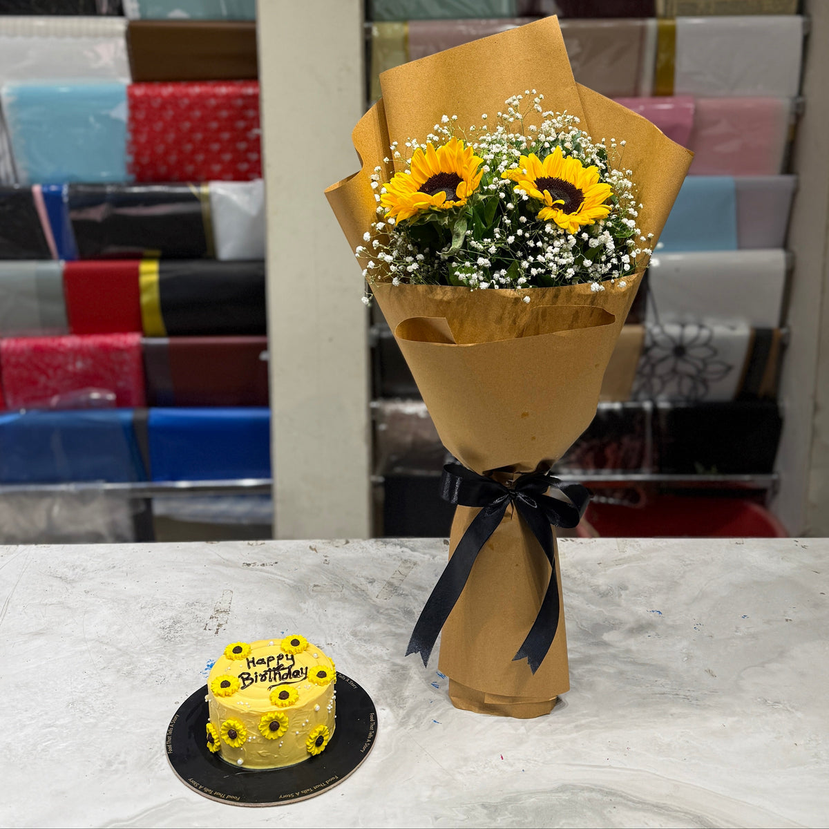A bouquet of sunflower and baby's breath flowers wrapped in brown paper with a black ribbon, placed on a marble counter next to a small yellow sunflower cake, with a colorful gift shop background.