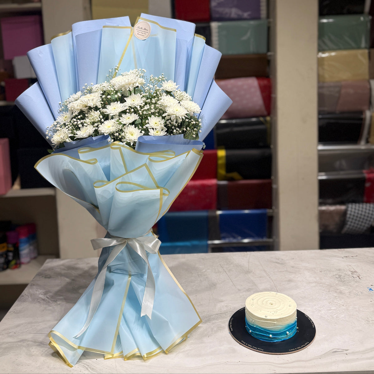 A light blue and gold wrapped flower bouquet with white flowers sits on a marble counter next to a small round cake in a gift shop setting with shelves of colorful wrapping paper in the background.