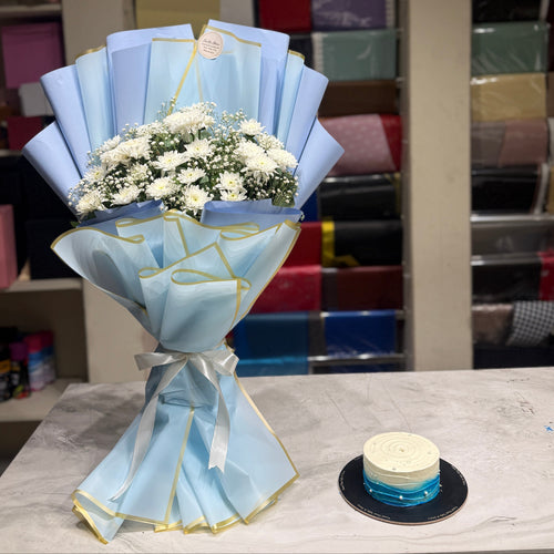 A light blue and gold wrapped flower bouquet with white flowers sits on a marble counter next to a small round cake in a gift shop setting with shelves of colorful wrapping paper in the background.