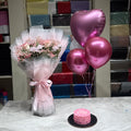 A pink-themed birthday gift set displayed on a marble table, featuring a bouquet of pink roses in white tulle, three metallic pink heart-shaped balloons, and a small pink frosted cake. The background shows shelves filled with colorful gift wrap rolls.
