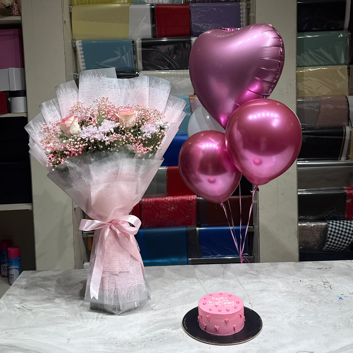 A pink-themed birthday gift set displayed on a marble table, featuring a bouquet of pink roses in white tulle, three metallic pink heart-shaped balloons, and a small pink frosted cake. The background shows shelves filled with colorful gift wrap rolls.