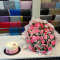 A large bouquet of pink and red flowers with white baby's breath wrapped in sheer pink paper sits on a white counter next to a small pink and white tiered cake. Behind them is a display rack filled with colorful gift boxes.