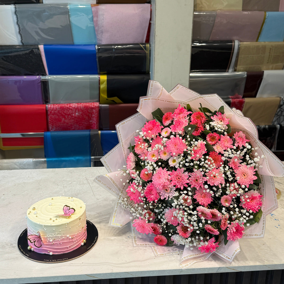 A large bouquet of pink and red flowers with white baby's breath wrapped in sheer pink paper sits on a white counter next to a small pink and white tiered cake. Behind them is a display rack filled with colorful gift boxes.