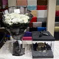 A bouquet of white flowers wrapped in black tissue paper and black cellophane stands next to a black gift box with a clear front panel and butterfly decoration on a marble-like surface. A colorful display of gift boxes is visible in the background.