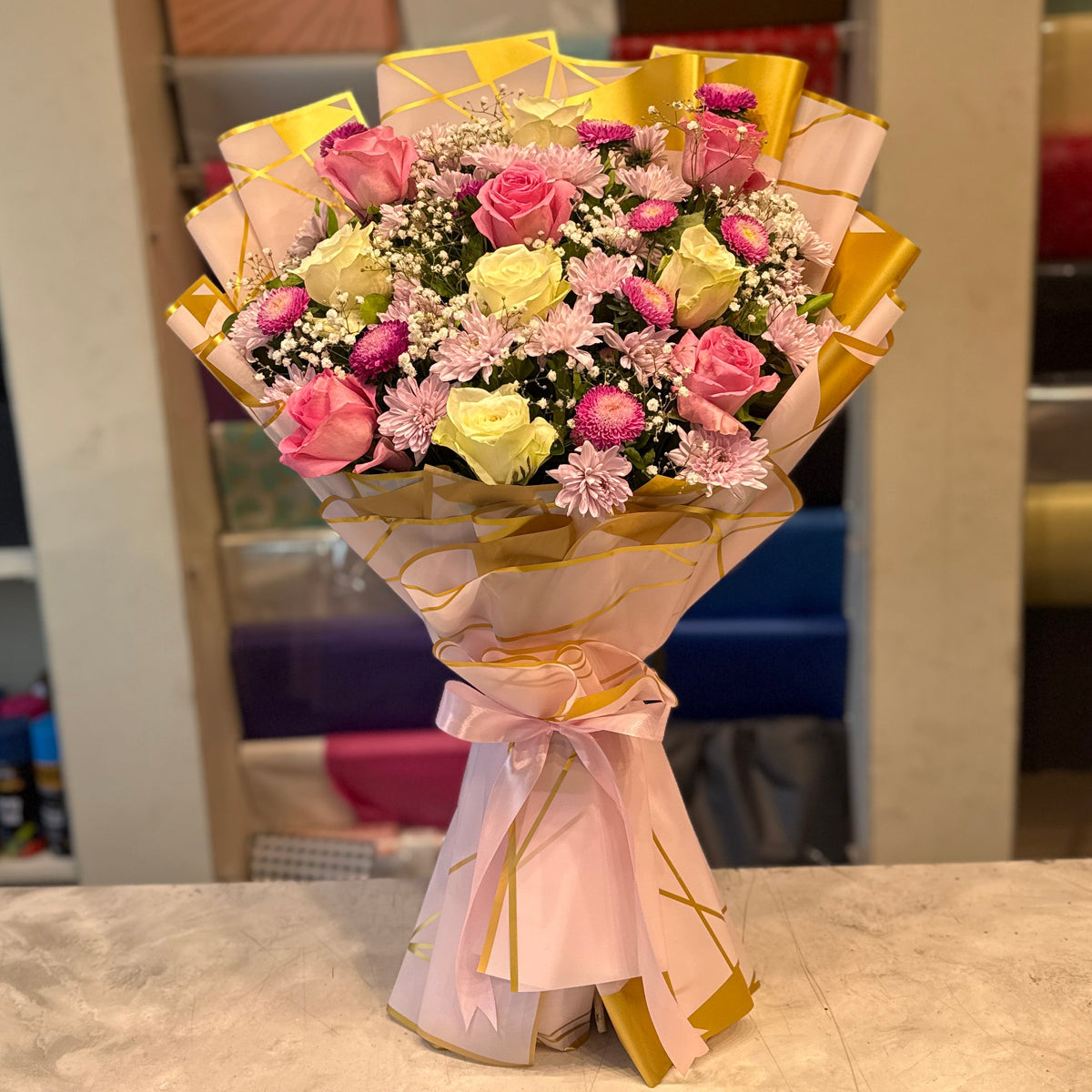 A vibrant bouquet of pink, yellow, and white roses wrapped in pink and gold paper, standing on a marble countertop with a blurred gift shop background.