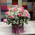 A floral arrangement of pink roses and white baby's breath in a pink gift box with a ribbon, displayed on a marble counter in front of a wall filled with colorful wrapped gift boxes.
