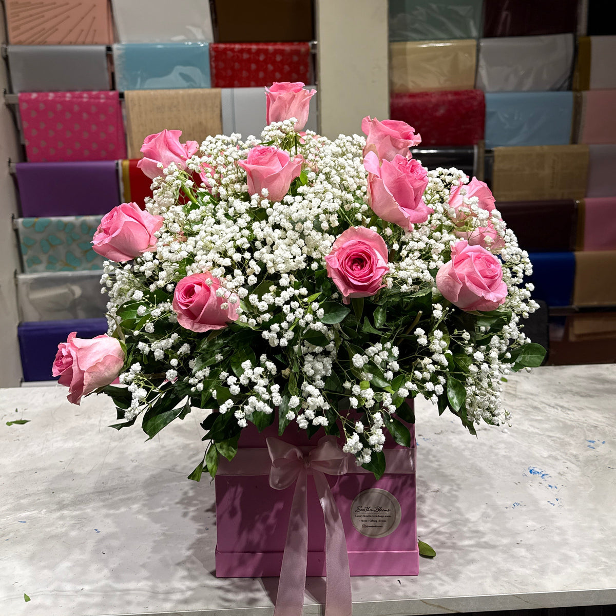 A floral arrangement of pink roses and white baby's breath in a pink gift box with a ribbon, displayed on a marble counter in front of a wall filled with colorful wrapped gift boxes.