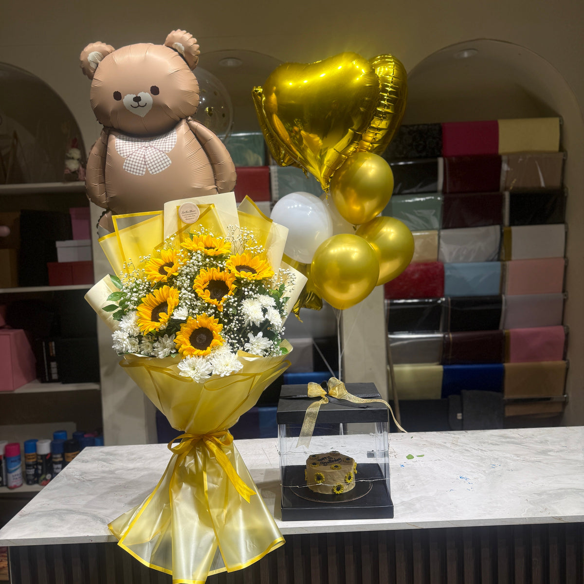 A gift set displayed on a marble counter, featuring a yellow-gold wrapped bouquet of sunflowers and white filler flowers, a small cake in a clear box, a brown teddy bear balloon, and gold foil heart and round balloons.
