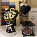 A black and yellow flower bouquet with white flowers and a black heart-shaped balloon next to a chocolate cake in a clear box on a marble table, with a shelf in the background containing folded fabrics and a teddy bear.