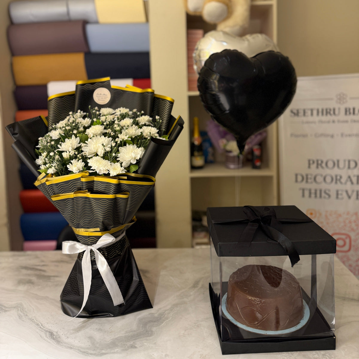 A black and yellow flower bouquet with white flowers and a black heart-shaped balloon next to a chocolate cake in a clear box on a marble table, with a shelf in the background containing folded fabrics and a teddy bear.