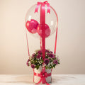 A decorative gift box arrangement on a marble-like surface, featuring a white cylindrical base filled with pink and purple flowers, tied with bright pink ribbon, and topped with a large clear balloon and smaller pink balloons in a shop interior.