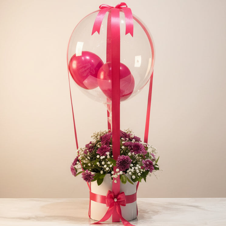 A decorative gift box arrangement on a marble-like surface, featuring a white cylindrical base filled with pink and purple flowers, tied with bright pink ribbon, and topped with a large clear balloon and smaller pink balloons in a shop interior.