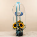 A decorative gift basket arrangement on a marble-like surface, featuring two large sunflowers, white baby's breath, and a clear balloon dome tied with a light blue ribbon. The background shows shelves with many wrapped gift boxes in different colors.