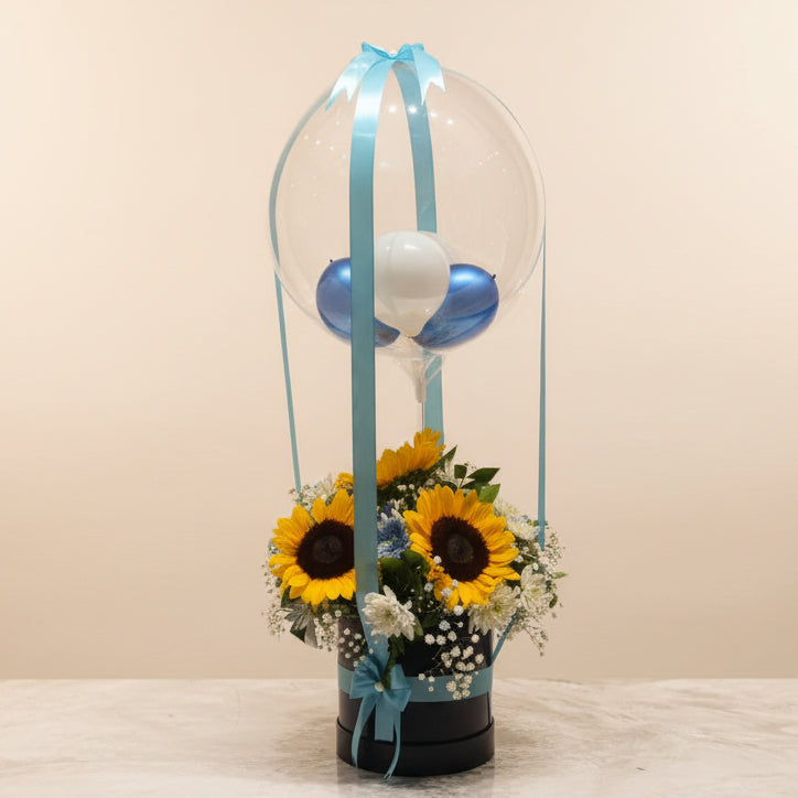 A decorative gift basket arrangement on a marble-like surface, featuring two large sunflowers, white baby's breath, and a clear balloon dome tied with a light blue ribbon. The background shows shelves with many wrapped gift boxes in different colors.