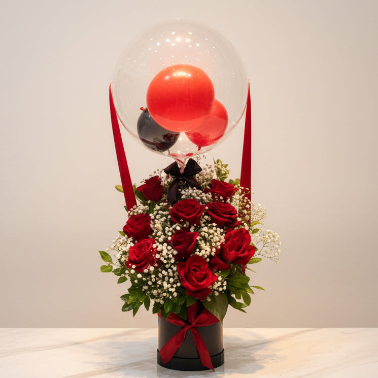 A floral centerpiece arrangement with red roses, baby's breath, and green foliage, topped with a clear balloon globe containing a red sphere, displayed on a table in front of a wall with shelves of colorful wrapped items.